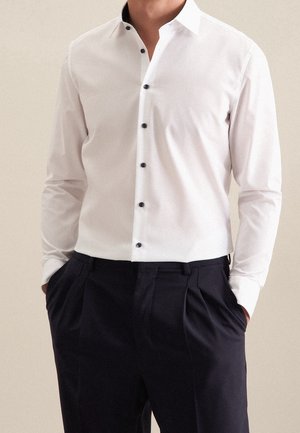 Mann trägt ein weißes langärmeliges Hemd mit schwarzen Knöpfen, in schwarze plissierte Hose gesteckt, die Hände in den Taschen.