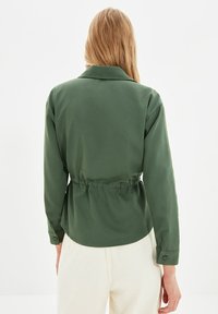 Vrouw met lang blond haar, gekleed in een aansluitende olijfgroene lange mouwenjas en witte broek, staand met haar rug naar de camera.