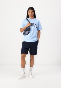 Světle modré bavlněné tričko s logem "LACOSTE", námořnické modré kraťasy a bílé tenisky. Doplňkem je černo-šedý batoh s efektem tie-dye.
