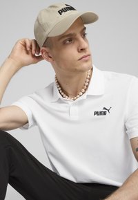 Polo bianca con logo Puma nero, indossata con un cappello beige. Una collana di perle aggiunge dettaglio. Il modello posa con una mano sulla testa. Pantaloni neri visibili.