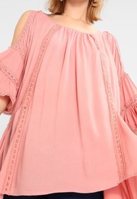 Růžový top s odkrytými rameny, s krajkovými detaily podél švů a rozšířenými rukávy, vyrobený z měkké, lehké tkaniny.