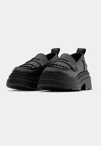 Svarte platåsko laget av glatt lær med en strukturert, mønstrete front og støtte i hælen. Inkluderer drasler for enkel på- og avkledning.