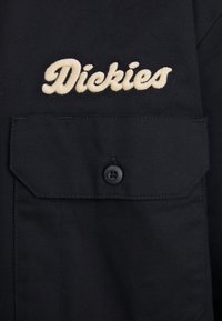 Camicia nera con bottoni e taschino sul petto, con logo "Dickies" ricamato in crema. Realizzata in un tessuto resistente, con una superficie liscia.