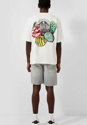 Mężczyzna w białej koszulce z kolorowymi owocami i napisem „Lemonade” na plecach, szare krótkie dżinsy, białe skarpetki i trampki.