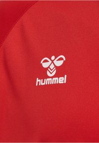 Weißes stilisiertes Hummel-Logo und „hummel“-Text auf rotem Stoffhintergrund, was auf Sportbekleidung oder athletische Kleidung hinweist.