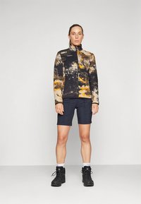 Ζακέτα από φλις με μοτίβο tie-dye σε μαύρο, μπεζ και πορτοκαλί, με ψηλό γιακά και φερμουάρ· συνδυασμένη με σκούρα σορτς και μαύρα ορειβατικά μποτάκια.