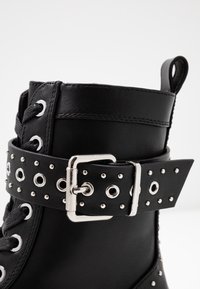 Bota de tobillo de cuero negro con ojales plateados, una tira negra con tachuelas y un hebilla metálica, y cordones negros. Textura suave y diseño resistente.