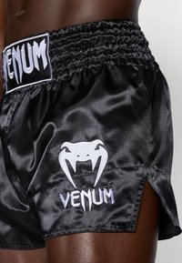 Pantalones cortos de boxeo de satén negro con cinturilla elástica fruncida, que presentan un logo blanco de "Venum" y un diseño de araña bordado.