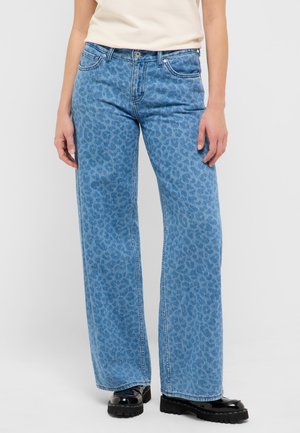 Osoba nosząca wysokie, szerokie niebieskie dżinsy z subtelnym wzorem w panterkę oraz czarne masywne buty, stojąca na gładkim tle.