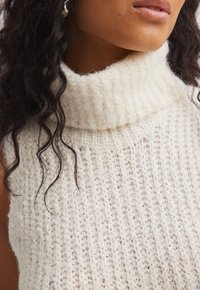 Chunky krem turtleneck pulover, izdelan iz teksturiranega pletenega materiala, s prostornim krojem in diskretnimi bež pikami po celotni tkanini.