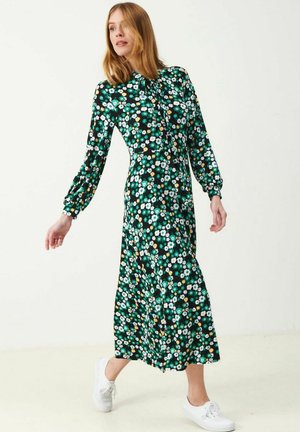 Nainen kävelee pitkähihaisessa, nilkkoihin ulottuvassa vihreässä mekossa, jossa on valkoinen ja keltainen kukkakuosi, yhdistettynä valkoisiin lenkkareihin yksiväristä taustaa vasten.