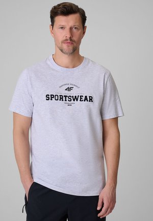 Mężczyzna ubrany w jasnoszarą koszulkę z krótkim rękawem z napisem „Sportswear” stojący na jednolitym szarym tle.