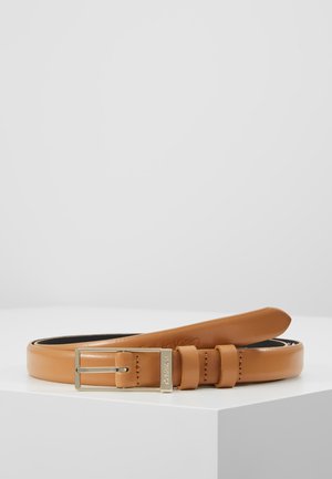Ceinture en cuir marron clair enroulée avec boucle argentée, exposée sur une surface blanche sur fond gris uni.
