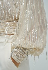 Durchsichtiger beiger Ärmeln mit silbernen Pailletten, gerafftem elastischem Bündchen mit Knöpfen, Hand in der hellbeigen Taschen des Kleidungsstücks gesteckt.