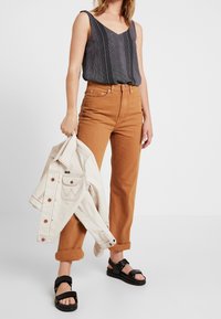 Donna che indossa un top senza maniche nero a fantasia, pantaloni marroni arrotolati, sandali neri, e tiene in una mano una giacca di jeans beige chiaro.