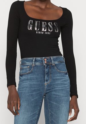 Person iført en sort langærmet top med teksten "GUESS Since 1981" i sølv og højtaljede blå jeans med dobbelte knapper.