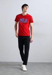 Piros póló "POLO" logóval kék színben, fekete nadrággal és fehér tornacipővel párosítva. Simára megmunkált anyag, rövid ujjú és kerek nyakú design.