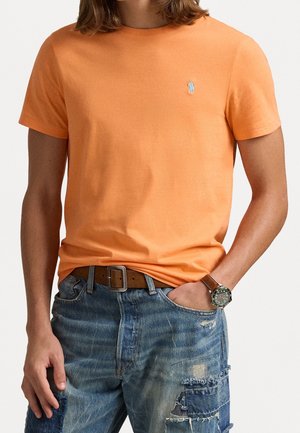 Pomarańczowa bawełniana koszulka z krótkimi rękawami, z małym niebieskim logo. Połączona z postrzępionymi niebieskimi dżinsami i brązowym paskiem.