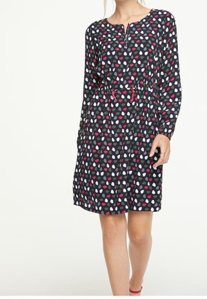 Femme portant une robe noire longueur genou avec des motifs gouttes multicolores, manches longues, fermeture éclair devant et taille resserrée par un cordon rose.