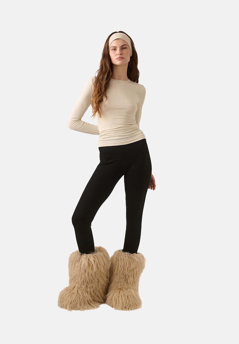 Bézs, hosszú ujjú felső fekete leggings-szel párosítva. A modellen plüss, világosbarna, szőrős csizma van, amely puha textúrával és széles nyílással rendelkezik a felső részén.