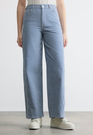 Személy, aki magasderekú, kék-fehér függőlegesen csíkozott, bő szárú nadrágot visel fehér platform tornacipővel, szürke padlón áll.