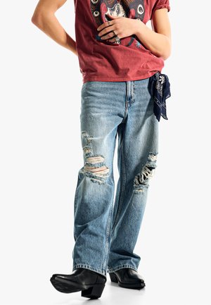 Osoba w podartych jasnoniebieskich dżinsach z czarną bandaną w kieszeni, czerwonym koszulce z nadrukiem i czarnych botkach na obcasie.