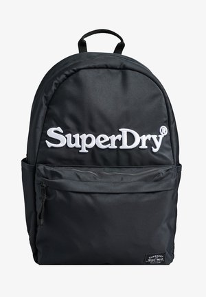 Zaino in tessuto nero con schienale imbottito, manico superiore, spallacci regolabili, tasca anteriore con zip e logo "SuperDry" bianco sul davanti.
