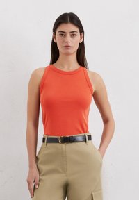 Canotta arancione brillante con scollo rotondo e design senza maniche, abbinata a pantaloni cargo beige e una cintura nera. Texture liscia, forma aderente.