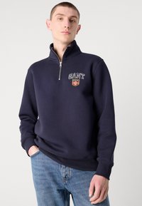Sweatshirt de marin cu fermoar până la jumătate și guler înalt, având logo-ul GANT și emblema scut pe piept, asortat cu blugi de un albastru deschis.