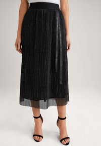 Černá plisovaná midi sukně s texturovaným leskem, širokým pasem a stužkovým detailem, kombinovaná s černými sandály na řemínky.