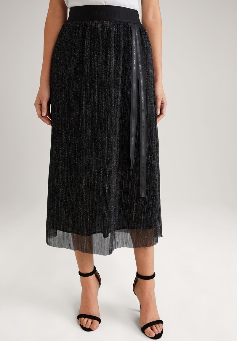 Černá plisovaná midi sukně s texturovaným leskem, širokým pasem a stužkovým detailem, kombinovaná s černými sandály na řemínky.