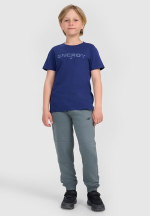 Dziecko stoi przodem, ubrane w granatowy t-shirt, szare dresowe spodnie z logo i czarne trampki na jednolitym jasnym tle.