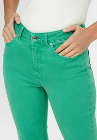 Green denim jeans feature a fitted design with front pockets, a silver button closure, and a smooth texture.