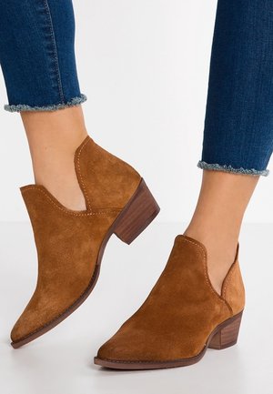 Braune Wildleder-Stiefeletten mit spitzen Zehen und niedrigen gestapelten Absätzen, getragen zu blauen Skinny-Jeans mit ausgefranstem Saum vor weißem Hintergrund.