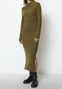 Robe en tricot côtelé vert olive avec des manches longues, un col montant et des fentes sur les côtés. Associée à des bottes à talons épais noires.