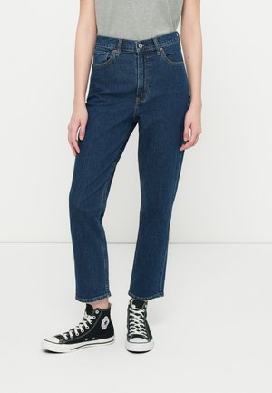 Personne portant un jean bleu droit coupé et des baskets montantes noires avec des lacets blancs, debout devant un fond blanc uni.