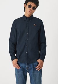 Cămașă albastru închis, cu mâneci lungi, croială slim, având un mic design brodat pe piept. Accentuata cu butoane transparente.