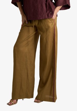 Persona che indossa pantaloni larghi color senape con vita con coulisse e un top bordeaux, in piedi con una mano in tasca e sandali dorati con tacco.