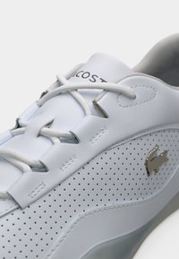 Baskets blanches perforées avec lacets blancs, coutures courbées et petit logo crocodile argenté sur le côté.