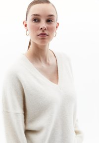 Maglione oversize a V in morbido tessuto bianco con una superficie liscia. La modella indossa orecchini a cerchio dorati, sullo sfondo neutro.
