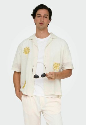 Short-sleeve, light beige shirt with yellow floral embroidery, buttoned front, and relaxed fit, worn with a white t-shirt underneath.