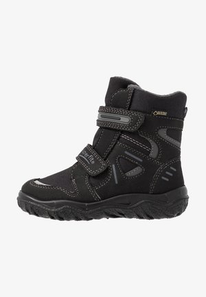 Bota preta impermeável com parte superior sintética, sola de borracha, bico redondo, tira de Velcro e detalhes refletivos para visibilidade em baixa luminosidade.