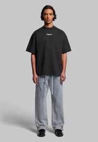 Black oversized t-shirt with short sleeves and crew neck, featuring white logo text on the chest, paired with light blue loose-fitting jeans.