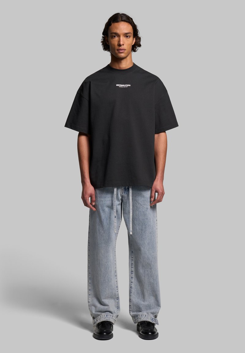 Black oversized t-shirt with short sleeves and crew neck, featuring white logo text on the chest, paired with light blue loose-fitting jeans.