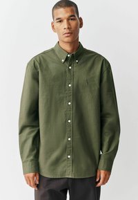 Chemise boutonnée vert olive en tissu doux et texturé. Caractéristiques : col pointu, manches longues et boutons blancs. Petite logo sur la poitrine.