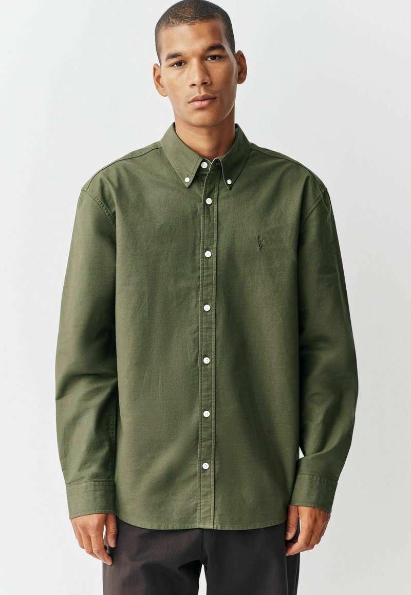 Chemise boutonnée vert olive en tissu doux et texturé. Caractéristiques : col pointu, manches longues et boutons blancs. Petite logo sur la poitrine.