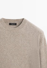Maglione in lana beige chiaro con collo rotondo a costine. Il tessuto ha una morbida trama con un leggero motivo variegato. Etichetta del marchio visibile.