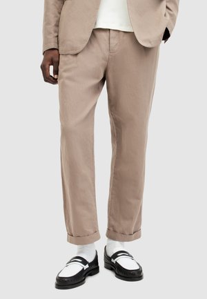 Osoba ubrana w beżowe spodnie garniturowe podwinięte przy kostkach, białe skarpetki, czarno-białe mokasyny oraz beżową marynarkę na białej bluzce, stojąca na tle szarego tła.