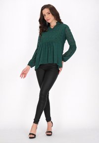 Grøn, semi-gennemsigtig bluse med teksturerede cirkulære accenter, V-hals og lange ærmer; parret med sorte faux læder leggings og sorte hæle.