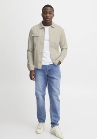 Light beige button-up jacket over a white t-shirt, paired with blue jeans and cream sneakers. Jacket has two front pockets and a relaxed fit.